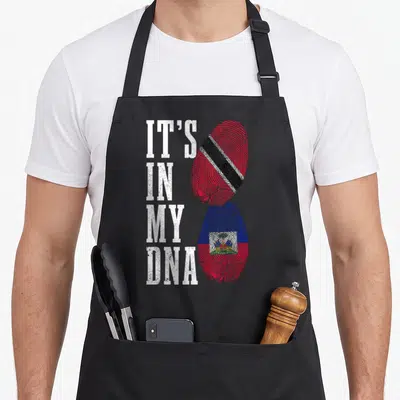 Haiti trinidad pride apron - IT'S IN MY DNA text next to red blue and black fingerprint flags representing Haiti — shown on a black cotton canvas apron against a white background