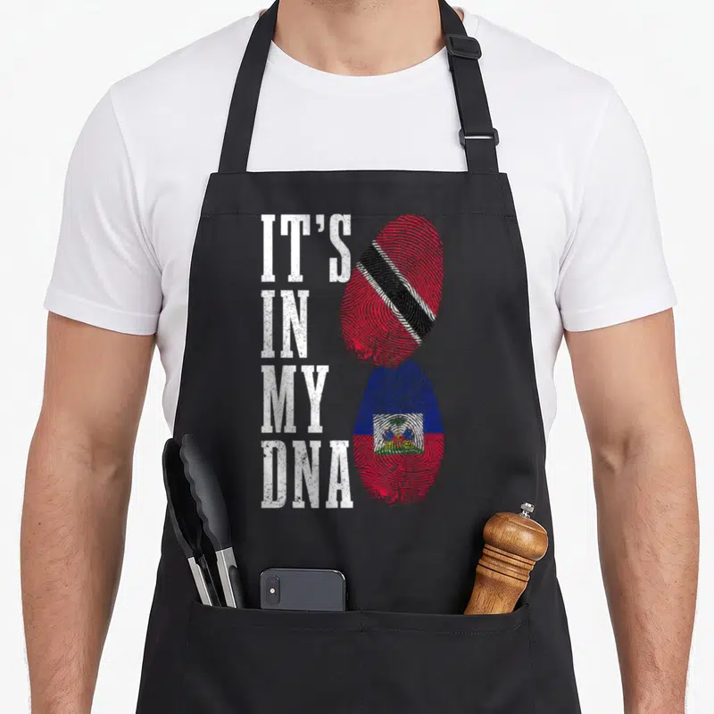 Haiti trinidad pride apron - IT'S IN MY DNA text next to red blue and black fingerprint flags representing Haiti — shown on a black cotton canvas apron against a white background