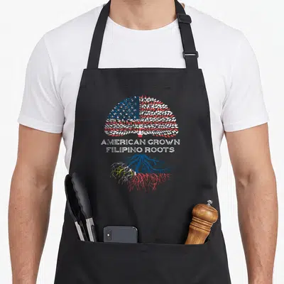 Filipino grown american flag apron - Distressed tree with flag-colored canopy and roots, stacked text AMERICAN GROWN — shown on a black cotton canvas apron against a white background