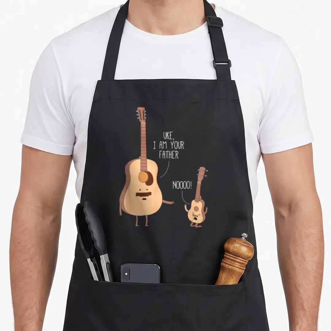 Uke fathers day apron - Acoustic guitar tells ukulele I am your father in cartoon style — shown on a black cotton canvas apron against a white background
