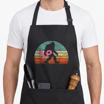 Breast bigfoot apron - Grey Bigfoot silhouette walking across retro striped sun with pink ribbon draped across chest — shown on a black cotton canvas apron against a white background