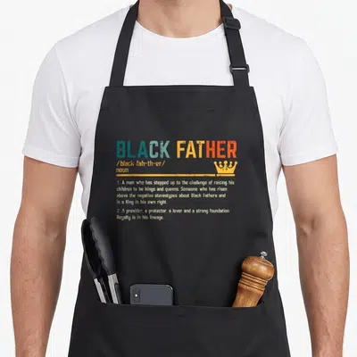 Definition african father day apron - Dictionary style text reading BLACK FATHER with crown icon in blue and orange — shown on a black cotton canvas apron against a white background