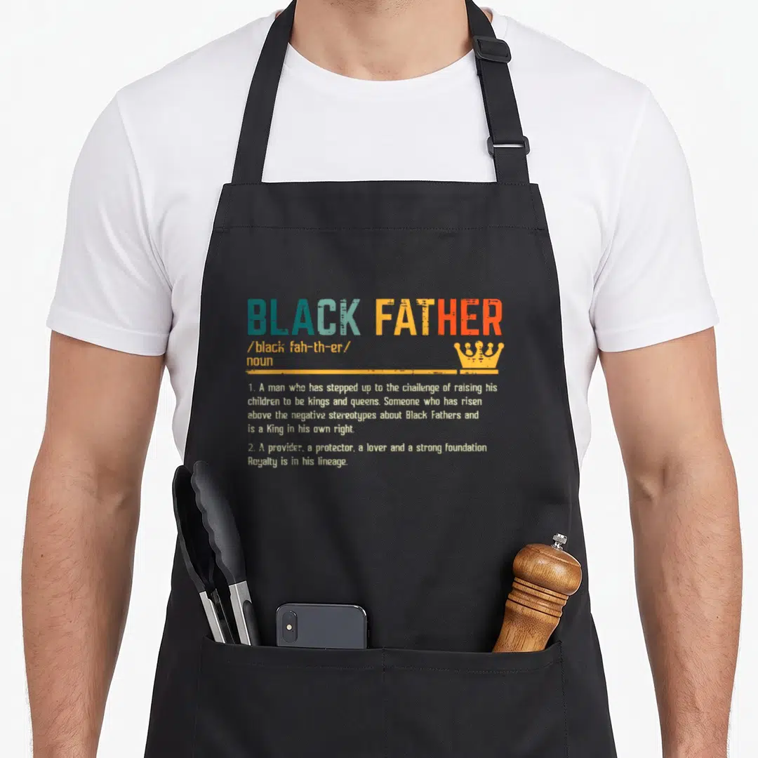 Definition african father day apron - Dictionary style text reading BLACK FATHER with crown icon in blue and orange — shown on a black cotton canvas apron against a white background
