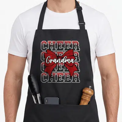 Bow grandma apron - Stacked CHEER text with red bow and Grandma script in red black white — shown on a black cotton canvas apron against a white background