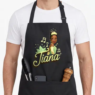 Tiana frog princess apron - Cartoon Tiana portrait above script text with musical notes and lily pad in green, yellow — shown on a black cotton canvas apron against a white background