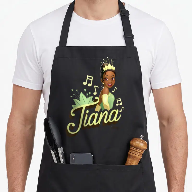 Tiana frog princess apron - Cartoon Tiana portrait above script text with musical notes and lily pad in green, yellow — shown on a black cotton canvas apron against a white background