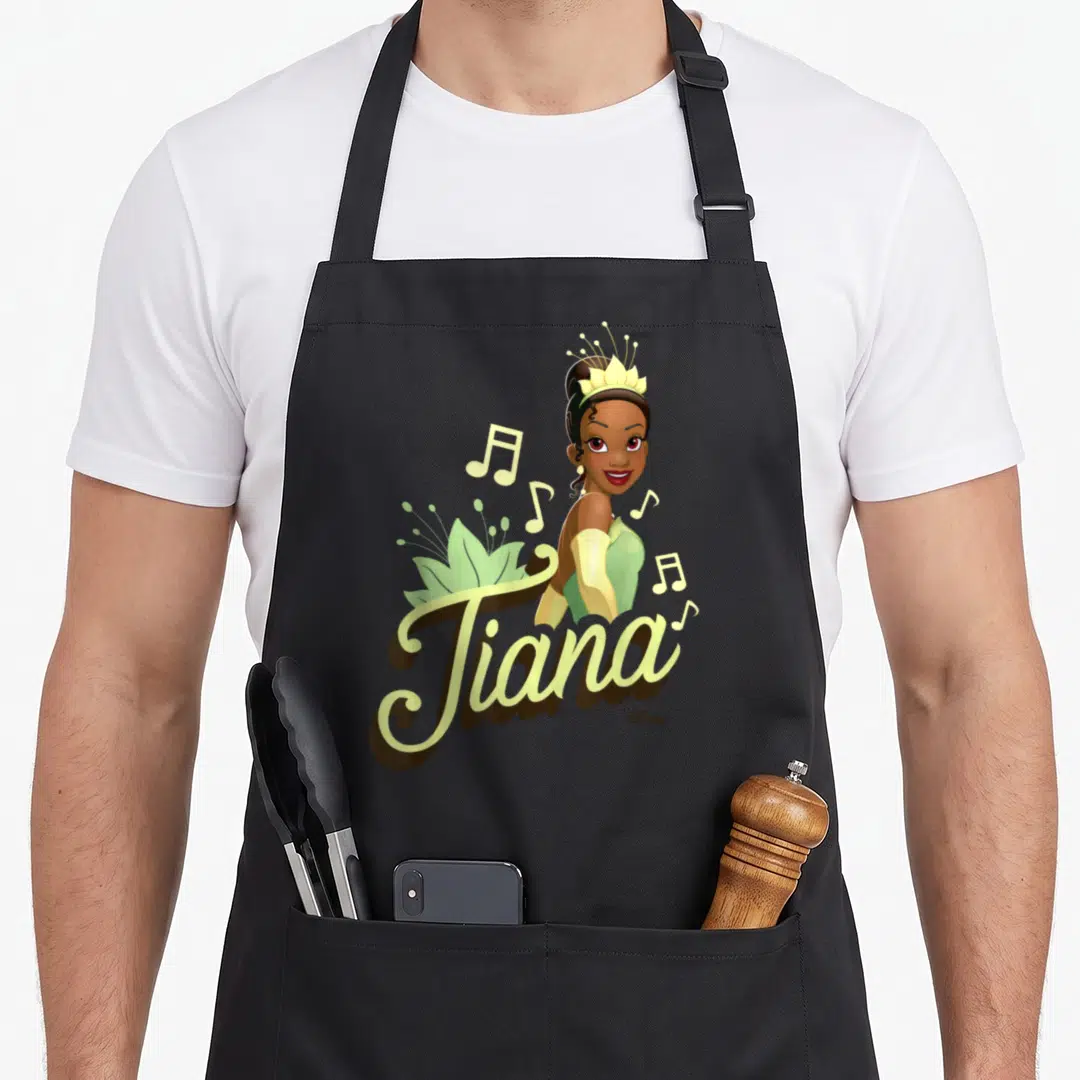 Tiana frog princess apron - Cartoon Tiana portrait above script text with musical notes and lily pad in green, yellow — shown on a black cotton canvas apron against a white background