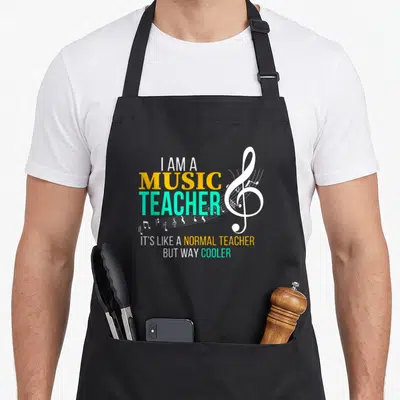 Funny music teacher apron - I AM A MUSIC TEACHER text with white treble clef and notes in yellow and turquoise — shown on a black cotton canvas apron against a white background