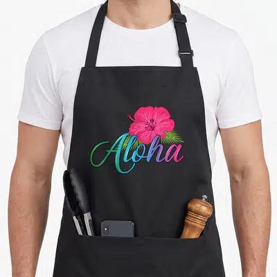 Aloha hawaii apron - Pink hibiscus flower over green palm leaf and gradient Aloha script text — shown on a black cotton canvas apron against a white background