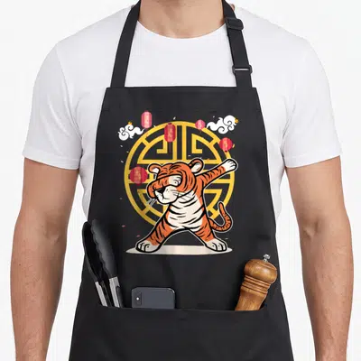 Dabbing new years eve apron - Cartoon tiger dabbing in front of yellow emblem with red lanterns and white clouds — shown on a black cotton canvas apron against a white background