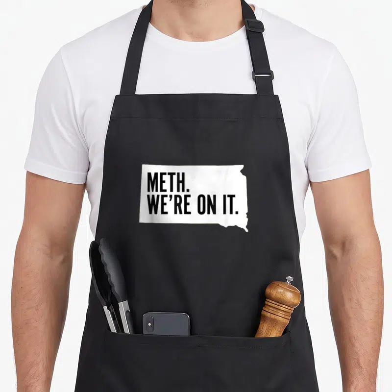 Meth meme apron - White North Dakota map shape containing black text METH. WE'RE ON IT — shown on a black cotton canvas apron against a white background