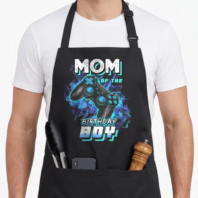 Born day video game apron - MOM OF THE BIRTHDAY BOY text surrounding a blue and purple cartoon game controller — shown on a black cotton canvas apron against a white background