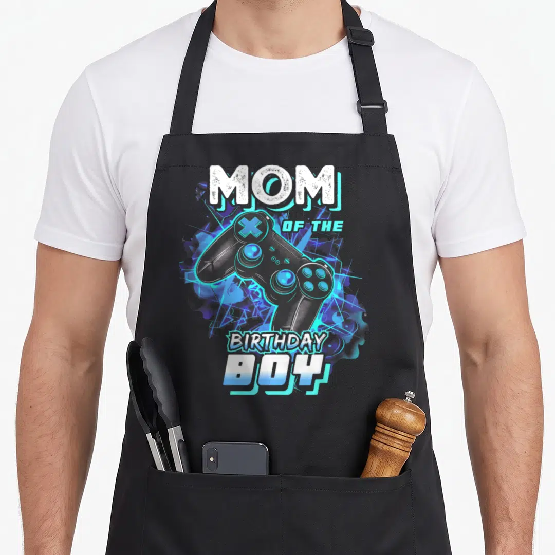 Born day video game apron - MOM OF THE BIRTHDAY BOY text surrounding a blue and purple cartoon game controller — shown on a black cotton canvas apron against a white background