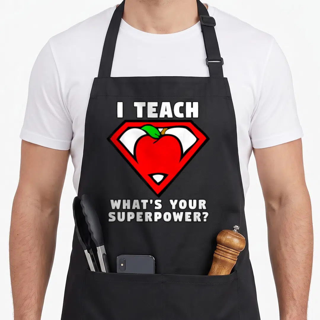 Teach apple apron - I Teach What's Your Superpower text with red shield and cartoon apple — shown on a black cotton canvas apron against a white background