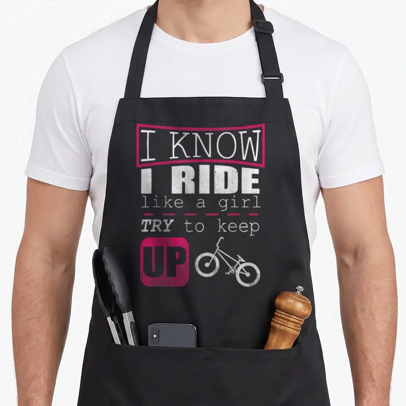 Ride bmx girl apron - Stacked white text I Know I Ride Like A Girl Try To Keep Up with pink accents and bicycle outline — shown on a black cotton canvas apron against a white background