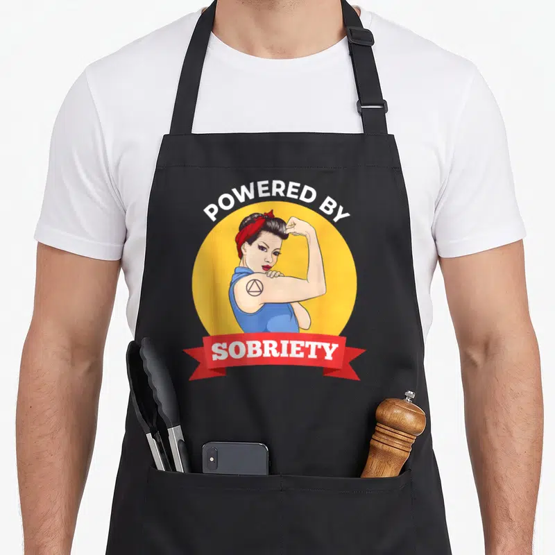 Sobriety coin vibes apron - Retro woman flexing arm with triangle tattoo inside yellow circle and POWERED BY SOBRIETY — shown on a black cotton canvas apron against a white background
