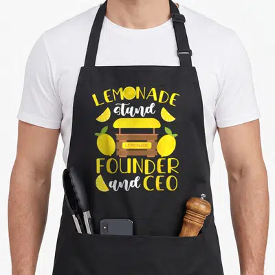 Ceo lemonade apron - Cartoon lemonade stand with yellow text LEMONADE stand FOUNDER and CEO surrounded by lemons — shown on a black cotton canvas apron against a white background