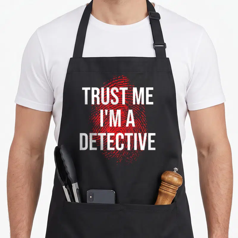 Trust detective apron - Red fingerprint graphic with white text TRUST ME I'M A DETECTIVE stacked vertically — shown on a black cotton canvas apron against a white background