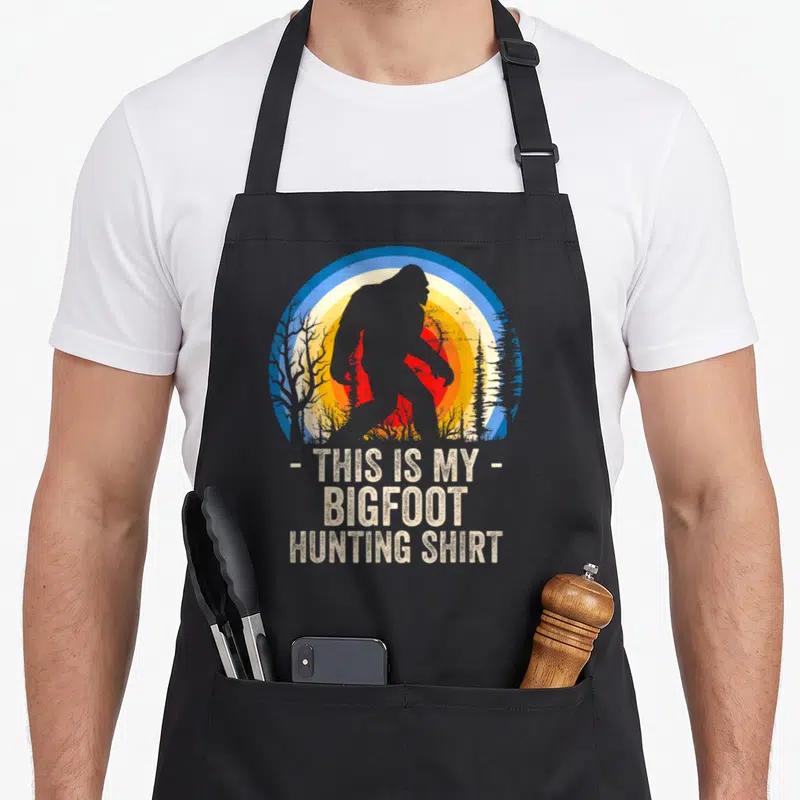 Hunting bigfoot apron - Retro Bigfoot silhouette over blue yellow red sunset with text THIS IS MY BIGFOOT HUNTING SHIRT — shown on a black cotton canvas apron against a white background