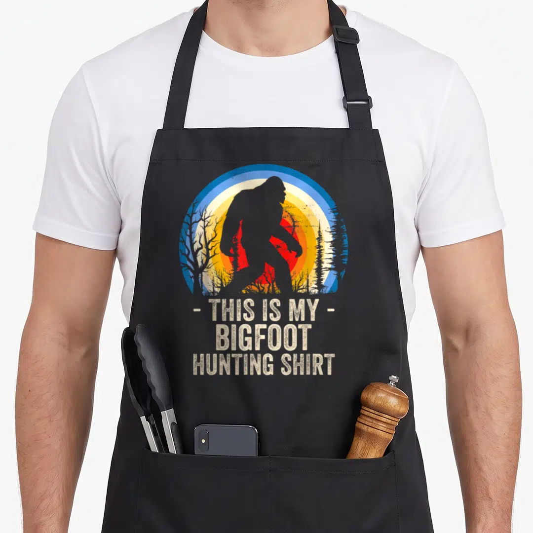 Hunting bigfoot apron - Retro Bigfoot silhouette over blue yellow red sunset with text THIS IS MY BIGFOOT HUNTING SHIRT — shown on a black cotton canvas apron against a white background