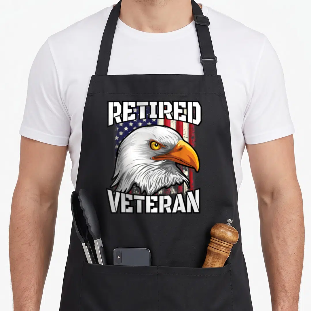 Retired eagles apron - Cartoon bald eagle head over distressed US flag with RETIRED VETERAN text in white red blue — shown on a black cotton canvas apron against a white background