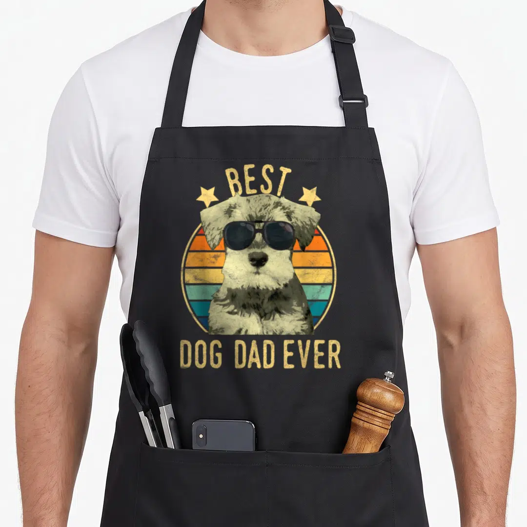 Father miniature schnauzer apron - Retro dog with sunglasses and striped sunset circle with text BEST DOG DAD EVER — shown on a black cotton canvas apron against a white background