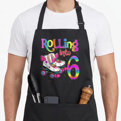 Rolling roll roller turning apron - Rolling into 6 rainbow text with white winged roller skates and stars — shown on a black cotton canvas apron against a white background