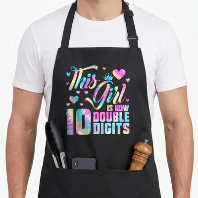 Birthday double apron - Tie-dye text This Girl Is Now Double Digits 10 with crown and hearts — shown on a black cotton canvas apron against a white background