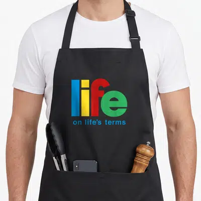 Term dealer funny apron - Stacked text life in blue yellow red green with on life's terms in blue below — shown on a black cotton canvas apron against a white background