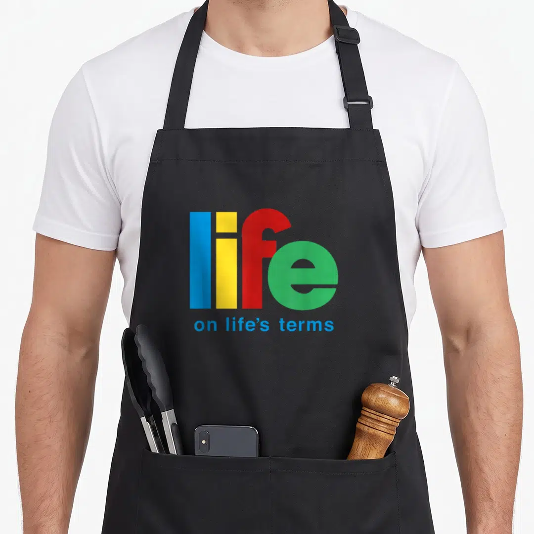 Term dealer funny apron - Stacked text life in blue yellow red green with on life's terms in blue below — shown on a black cotton canvas apron against a white background