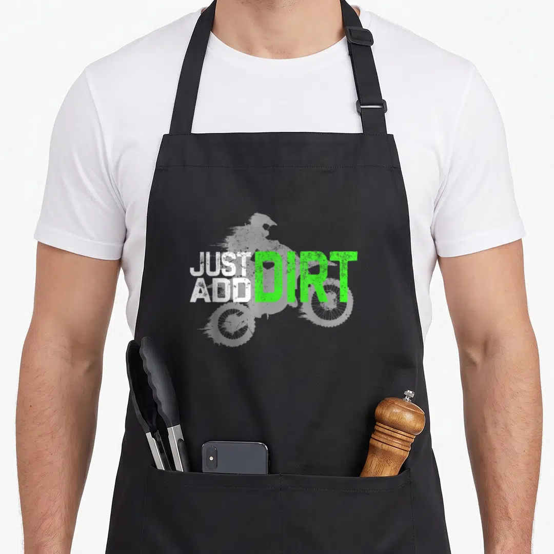 Youth bike apron - Grey dirt bike silhouette with stacked white text JUST ADD and large green text DIRT — shown on a black cotton canvas apron against a white background