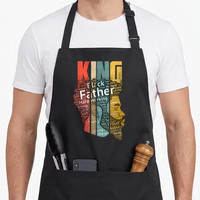 King african american aprons - KING text above three male profiles filled with words like Father, Son, Caring — shown on a black cotton canvas apron against a white background