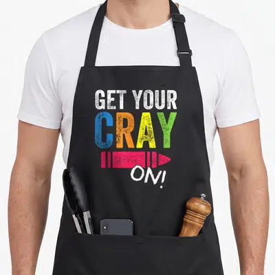 Funny cray teacher apron - GET YOUR CRAY ON! text with multi-colored letters and pink crayon drawing — shown on a black cotton canvas apron against a white background