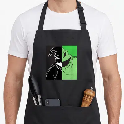 Oogie split apron - Split cartoon monster head in green and black with stitched details and two front pockets — shown on a black cotton canvas apron against a white background
