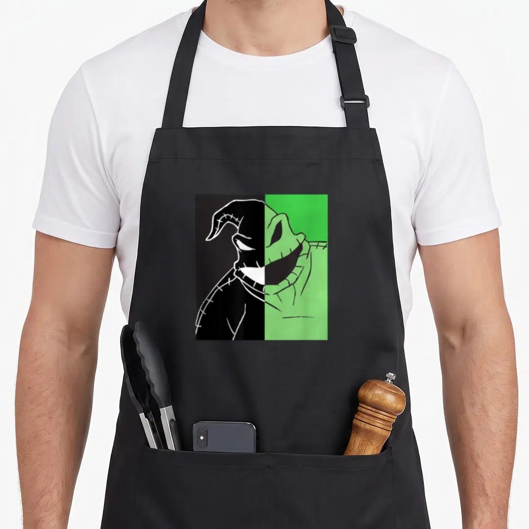 Oogie split apron - Split cartoon monster head in green and black with stitched details and two front pockets — shown on a black cotton canvas apron against a white background