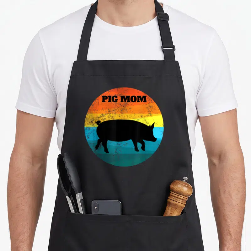 Pot pig lover apron - PIG MOM text over black pig silhouette on orange yellow blue teal retro sunset circle — shown on a black cotton canvas apron against a white background