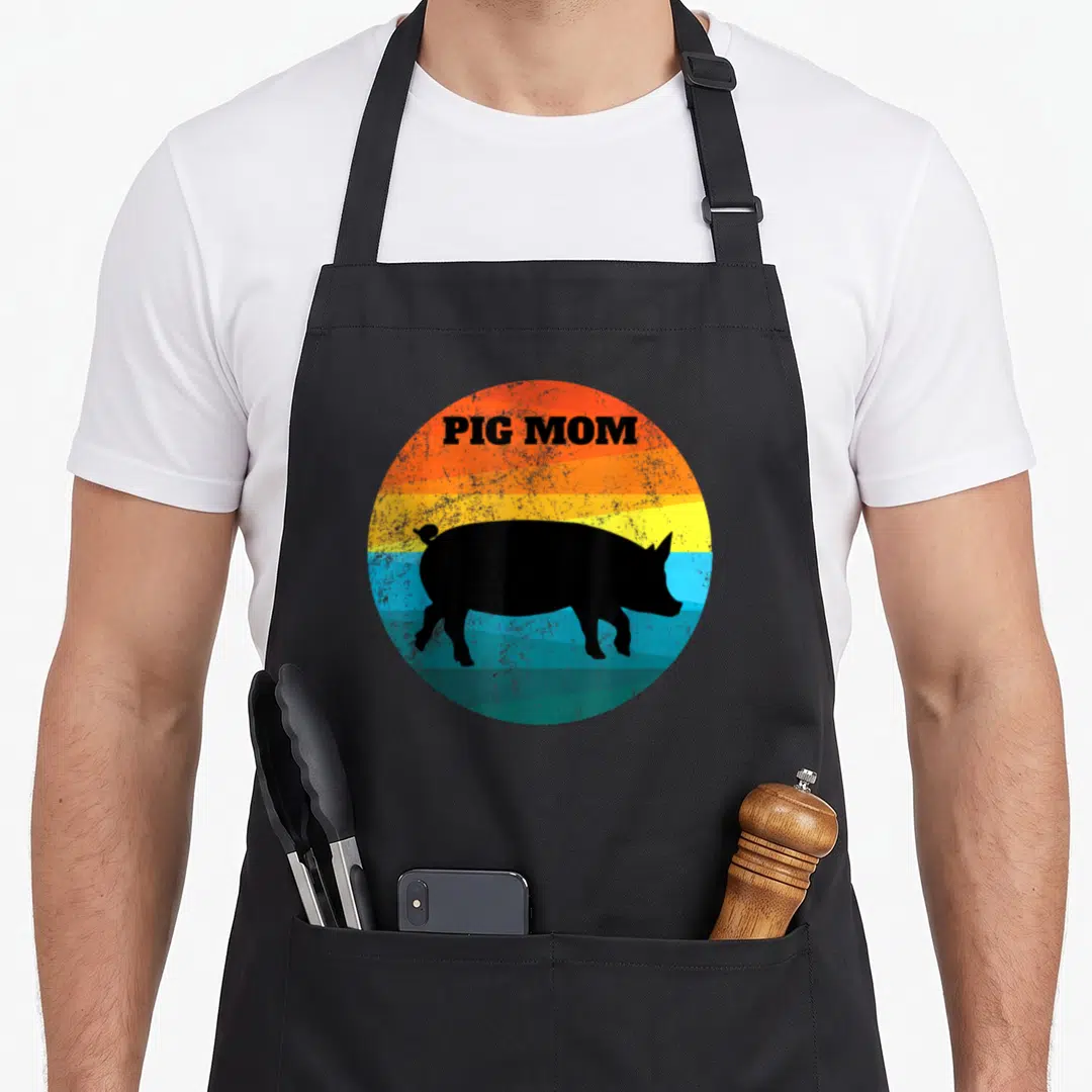 Pot pig lover apron - PIG MOM text over black pig silhouette on orange yellow blue teal retro sunset circle — shown on a black cotton canvas apron against a white background