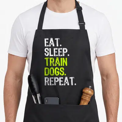 Eat dog training apron - Stacked white and lime green text reading EAT SLEEP TRAIN DOGS REPEAT — shown on a black cotton canvas apron against a white background