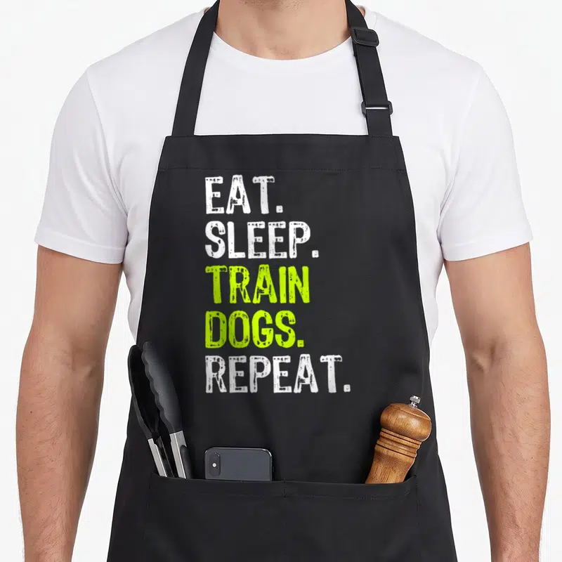Eat dog training apron - Stacked white and lime green text reading EAT SLEEP TRAIN DOGS REPEAT — shown on a black cotton canvas apron against a white background