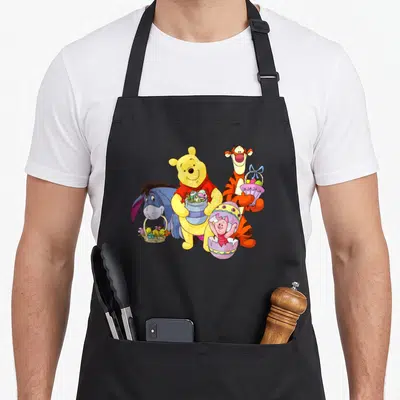 Character easter apron - Cartoon Winnie the Pooh holding eggs with Eeyore, Tigger, and Piglet in an Easter egg — shown on a black cotton canvas apron against a white background