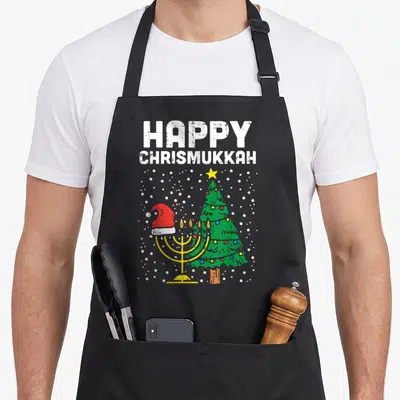 Christmukkah hanukkah apron - HAPPY CHRISMUKKAH text over yellow menorah in red santa hat beside green christmas tree — shown on a black cotton canvas apron against a white background