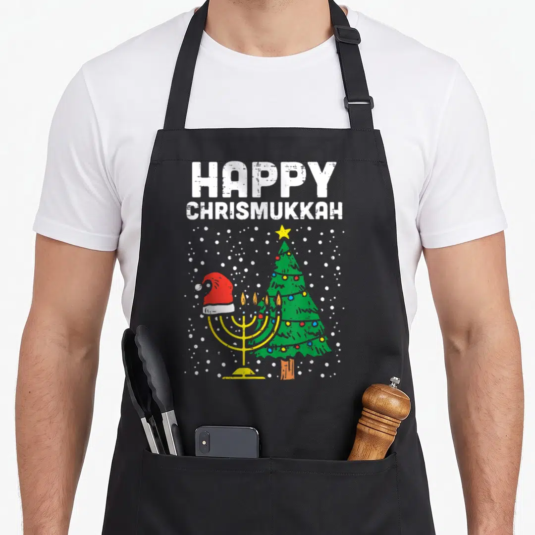 Christmukkah hanukkah apron - HAPPY CHRISMUKKAH text over yellow menorah in red santa hat beside green christmas tree — shown on a black cotton canvas apron against a white background