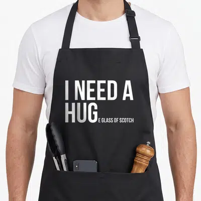 Huge whisky apron - White stacked text reading I NEED A HUG E GLASS OF SCOTCH pun — shown on a black cotton canvas apron against a white background