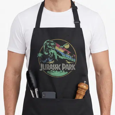 Darken novelty apron - Retro circular logo with green T-Rex, pterosaur, palm trees, and colorful diagonal stripes — shown on a black cotton canvas apron against a white background