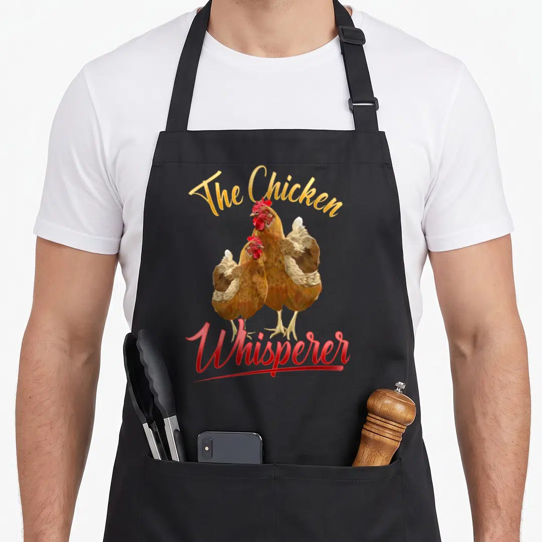 Farming chicken apron - Two brown chickens with red combs under gold The Chicken and red Whisperer text — shown on a black cotton canvas apron against a white background