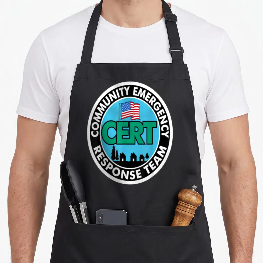 Cert community apron - Circular CERT logo with US flag, green text, and house silhouettes on a blue background — shown on a black cotton canvas apron against a white background