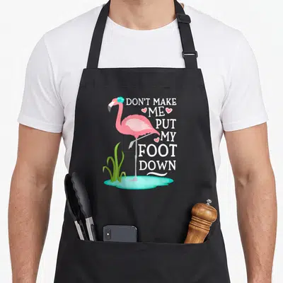 Cool foot pink flamingo apron - Cartoon pink flamingo in blue puddle with white stacked text and green grass accents — shown on a black cotton canvas apron against a white background