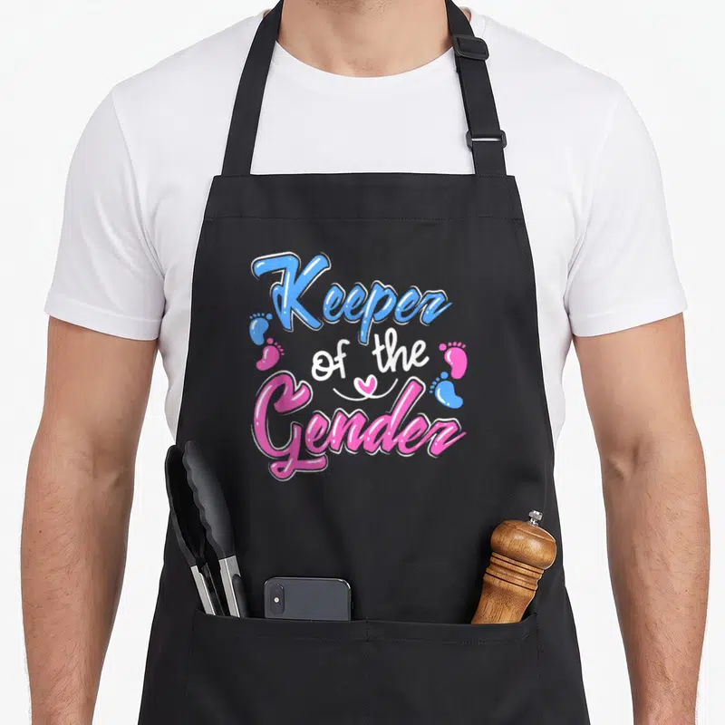 Announcement gender reveal apron - Blue pink and white text reading Keeper of the Gender with baby footprints and heart — shown on a black cotton canvas apron against a white background