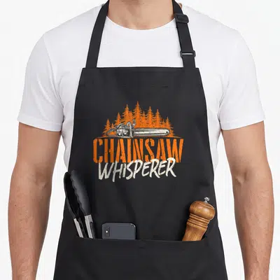 Chainsaw woodworker's apron - Orange pine trees behind a white chainsaw with stacked distressed text reading CHAINSAW — shown on a black cotton canvas apron against a white background