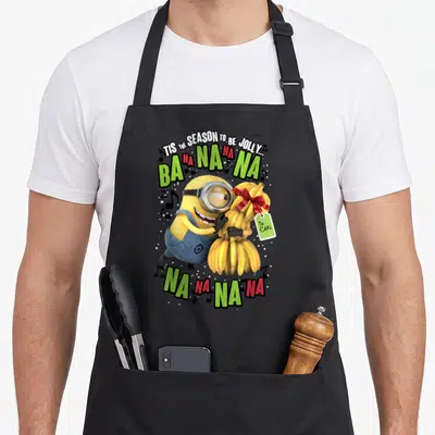 Despicable banana apron - Yellow Minion hugging bananas with red bow and green red text reading BA NA NA NA NA — shown on a black cotton canvas apron against a white background